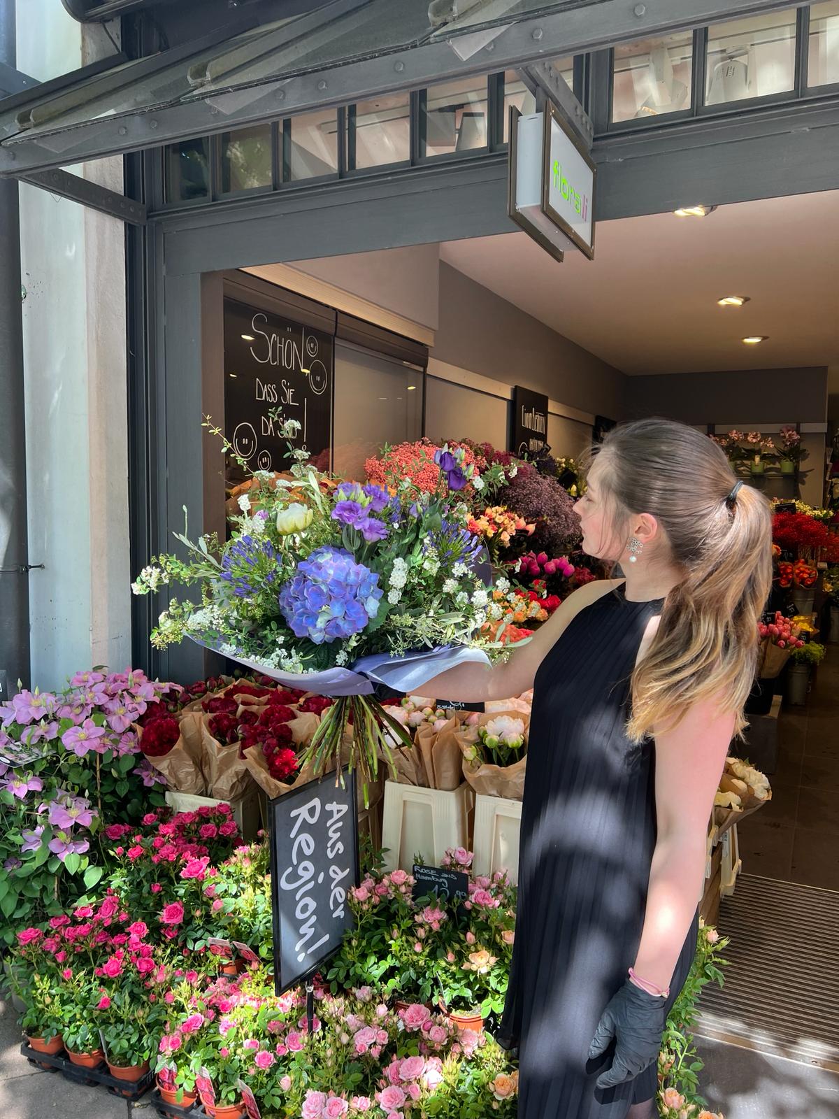 Eine Frau in einem schwarzen Kleid steht vor einem Blumenladen und hält einen großen Strauß bunter Blumen in der Hand. Hinter ihr sind verschiedene Blumenarrangements und Körbe im Ladeneingang zu sehen. Sonnenlicht fällt auf die Szene.