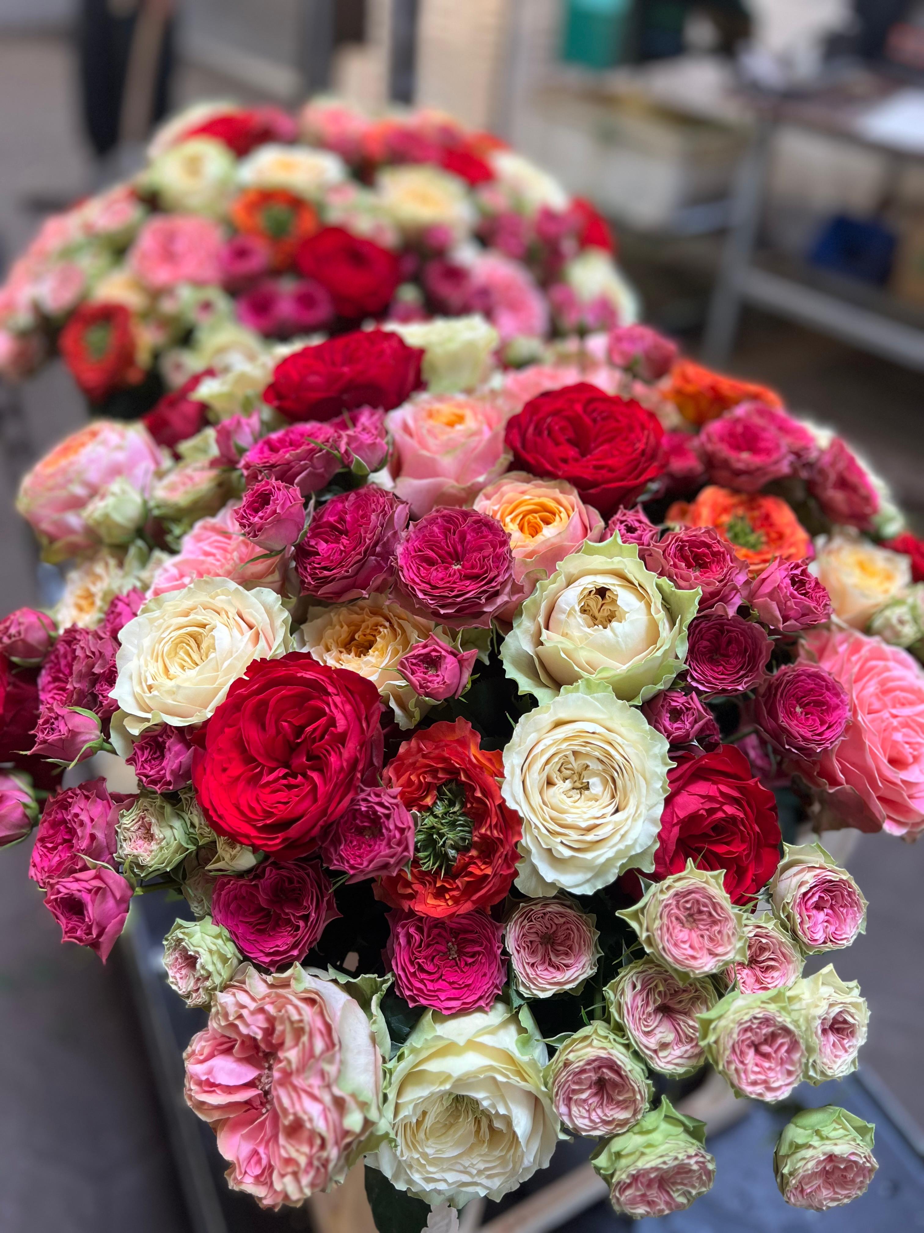 Ein großer Strauß verschiedener Rosen in verschiedenen Farben, darunter rot, rosa, weiß, pfirsichfarben und orange, die dicht nebeneinander auf einem Tisch in einem Blumenladen stehen.