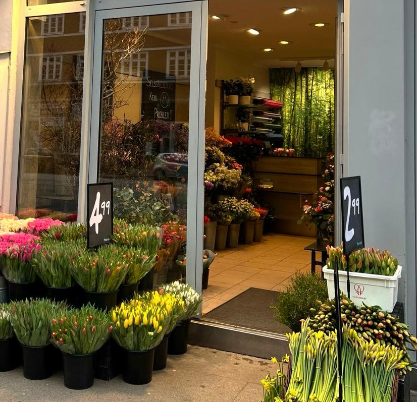 Der Eingang eines Blumenladens. Eimer mit Tulpen und verschiedene Blumen sind draußen ausgestellt. Zwei Preisschilder zeigen 4,99 und 2,99 an. Drinnen sind weitere Blumen an den Wänden und im hinteren Bereich arrangiert.