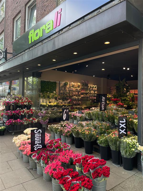 Ein Blumenladen namens Florali präsentiert vor seinem Eingang Eimer voller bunter Blumen, darunter Rosen und Nelken. Kreidetafeln zeigen Preise und Sonderangebote an. Die Schaufensterfront hat große Fenster und ein grünes Schild.