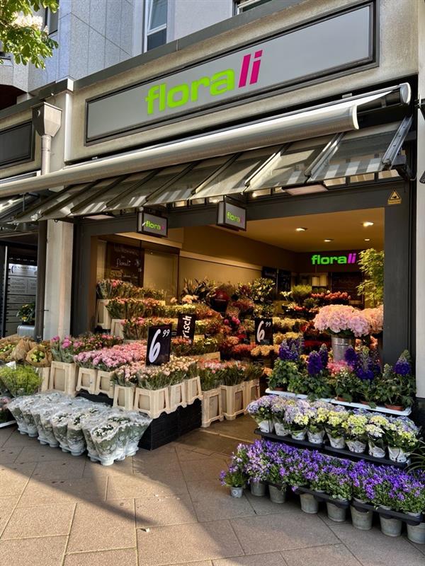 Ein Blumenladen namens „Florali“ präsentiert draußen und drinnen farbenfrohe Blumensträuße und Topfpflanzen, mit Preisschildern und verschiedenen Blumen, die ordentlich am und um den Eingang angeordnet sind.