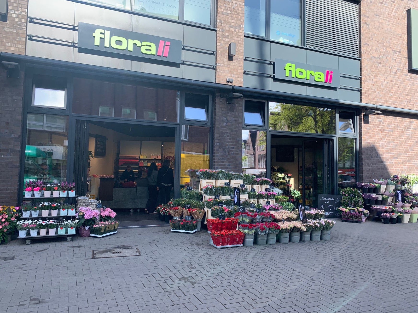 Ein Blumenladen namens Florali präsentiert vor seinem Eingang Eimer voller bunter Blumen, darunter Rosen und Nelken. Kreidetafeln zeigen Preise und Sonderangebote an. Die Schaufensterfront hat große Fenster und ein grünes Schild.