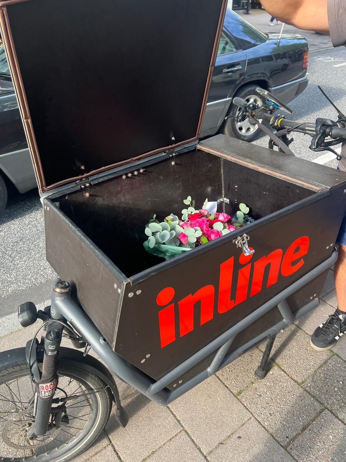 Ein schwarzes Lastenfahrrad mit einer großen, offenen Kiste, die in roten Buchstaben beschriftet ist, enthält einen kleinen Strauß Blumen und Grünzeug. Das Fahrrad ist auf einem Gehweg neben einer Straße und einem geparkten Auto abgestellt.