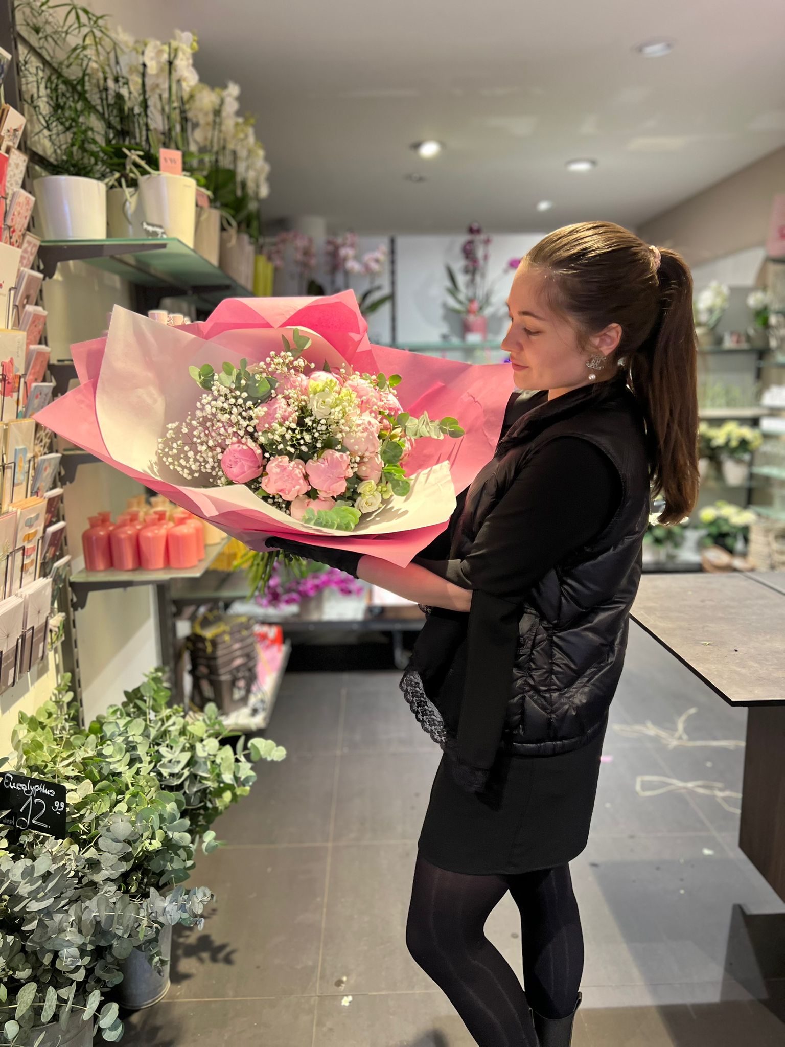 Eine Frau in einem schwarzen Kleid und einer Weste hält einen großen Strauß rosa und weißer Blumen, die in rosa Papier eingewickelt sind, und steht in einem Blumenladen voller Pflanzen, Blumen und Grußkarten.