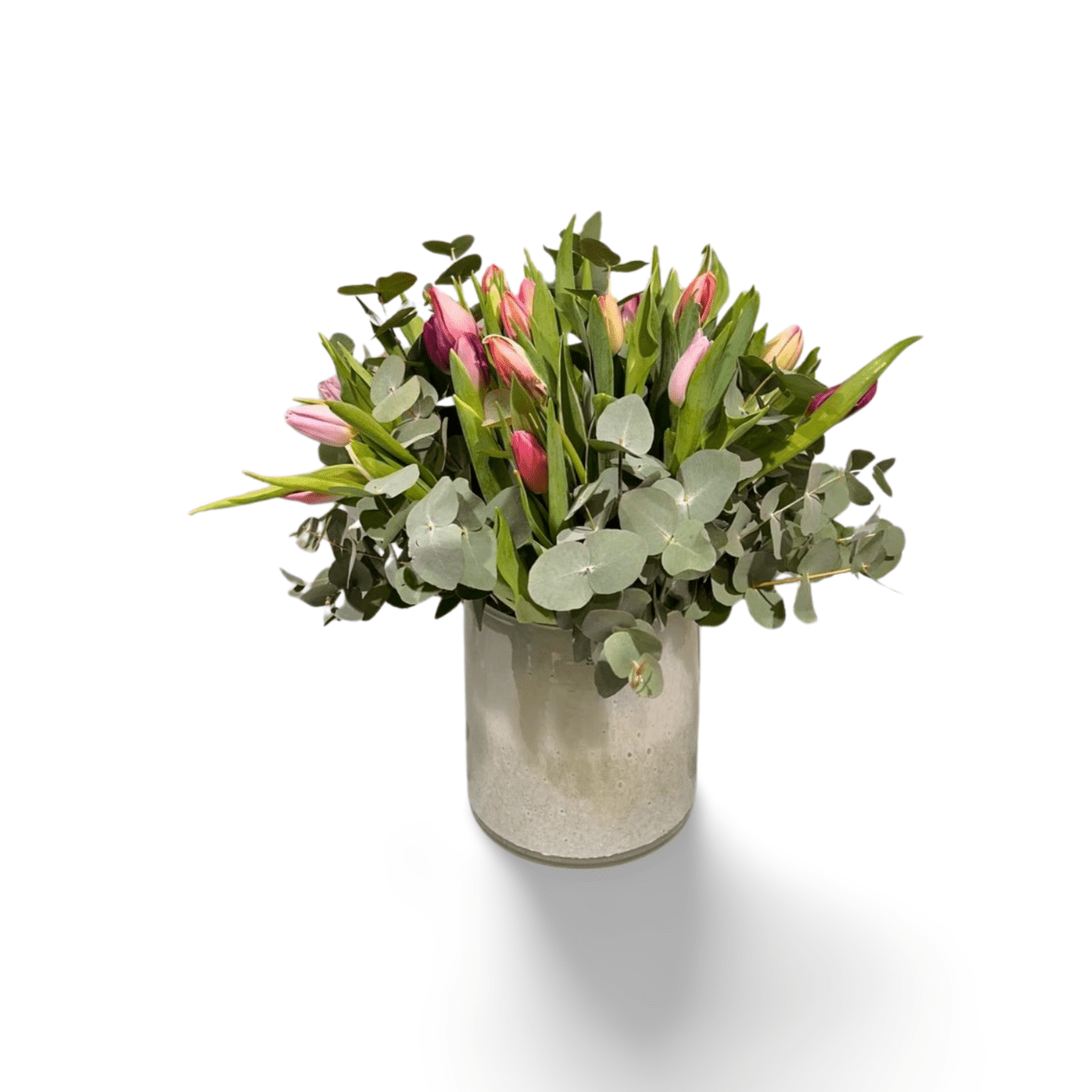 Der florali Tulip Dream besteht aus frischen rosa Tulpen und üppigen Eukalyptuszweigen in einer klaren Glasvase, wunderschön arrangiert vor einem weißen Hintergrund.