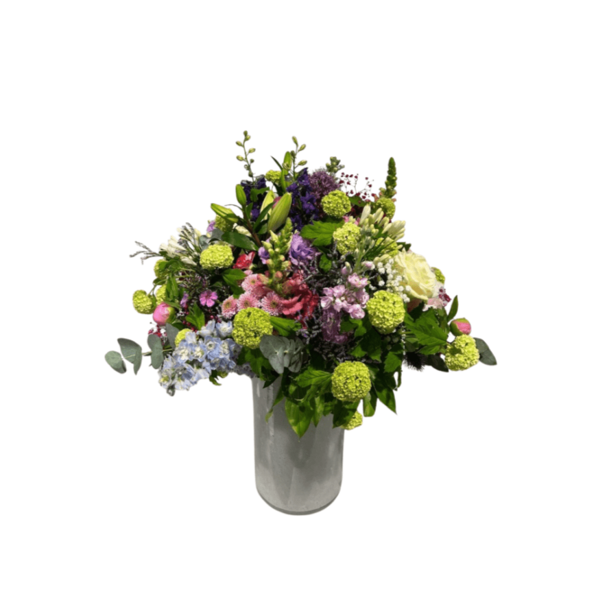 Das florali Wiesenspiel ist ein großer Strauß verschiedener violetter, rosafarbener, grüner und blauer Blumen, wunderschön arrangiert in einer weißen Vase vor einem schlichten weißen Hintergrund.