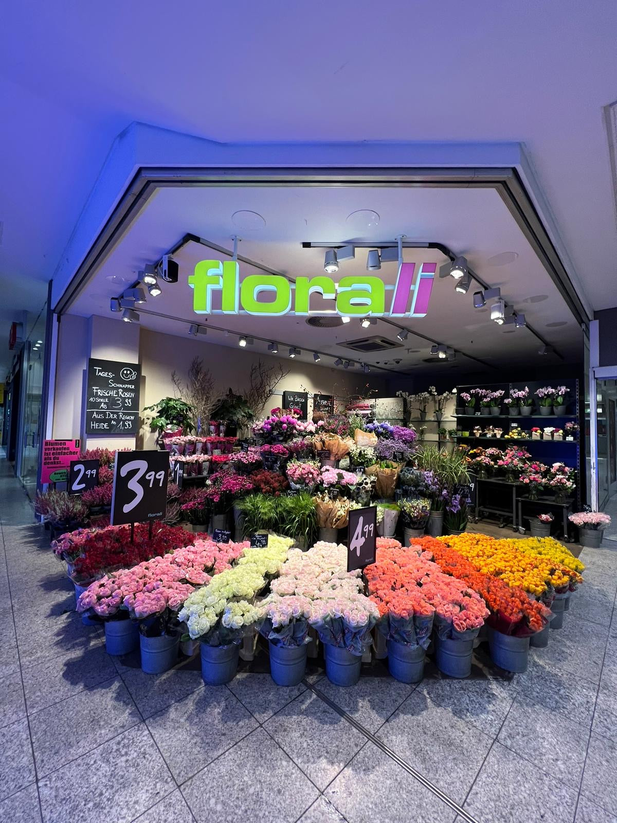 Ein Blumenladen namens Florali präsentiert vor dem Eingang eine Vielzahl farbenfroher frischer Blumen in Eimern mit Preisschildern. Drinnen sind unter hellem Licht weitere Blumen und Grünpflanzen an der Rückwand arrangiert.