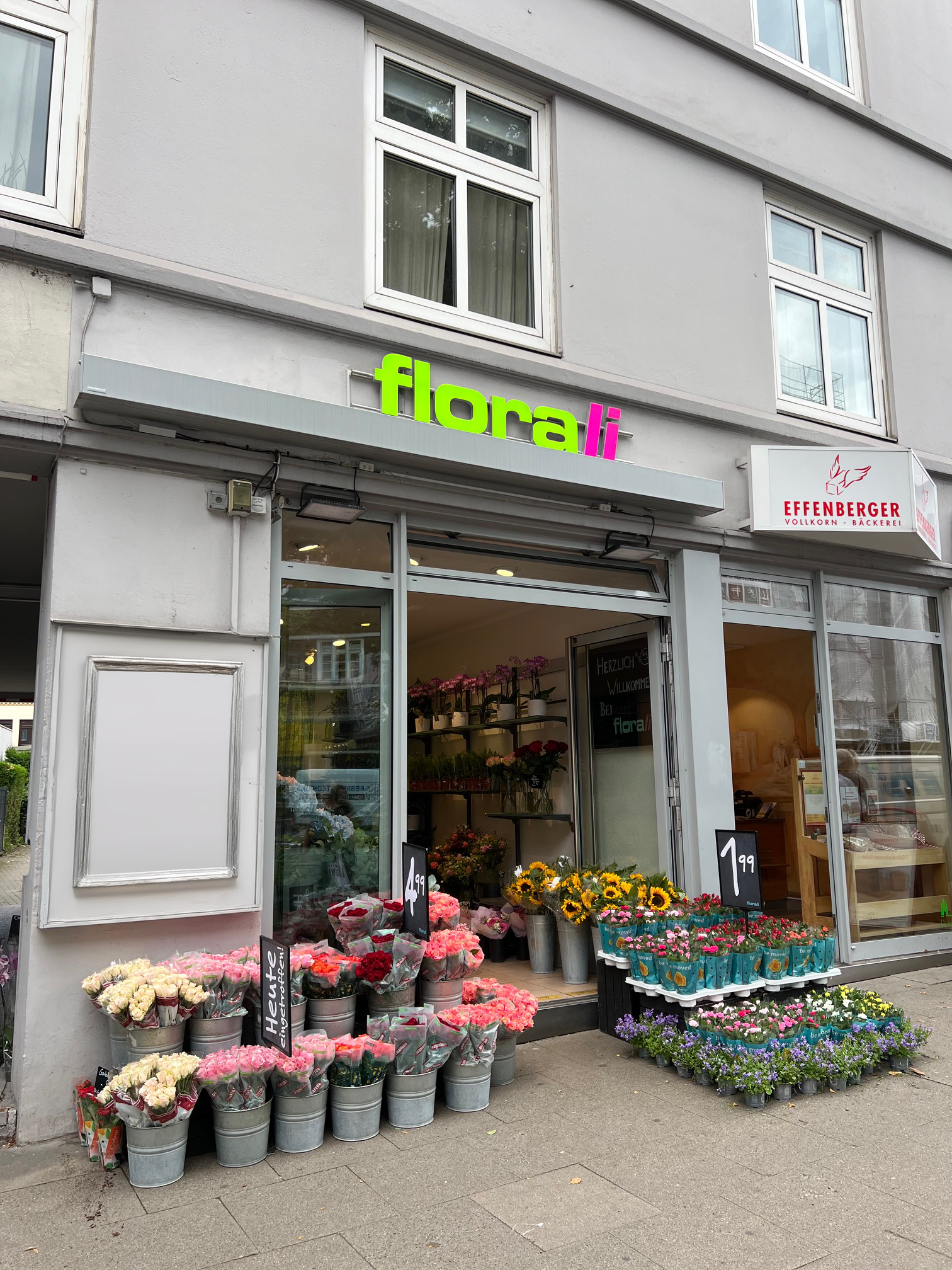 Ein Blumenladen namens Flora Li präsentiert bunte Blumen in Eimern und Schalen im Freien. Nebenan befindet sich die Bäckerei Effenberger. Beide Geschäfte haben große Fenster an einem grauen Gebäude mit Wohnungen darüber.