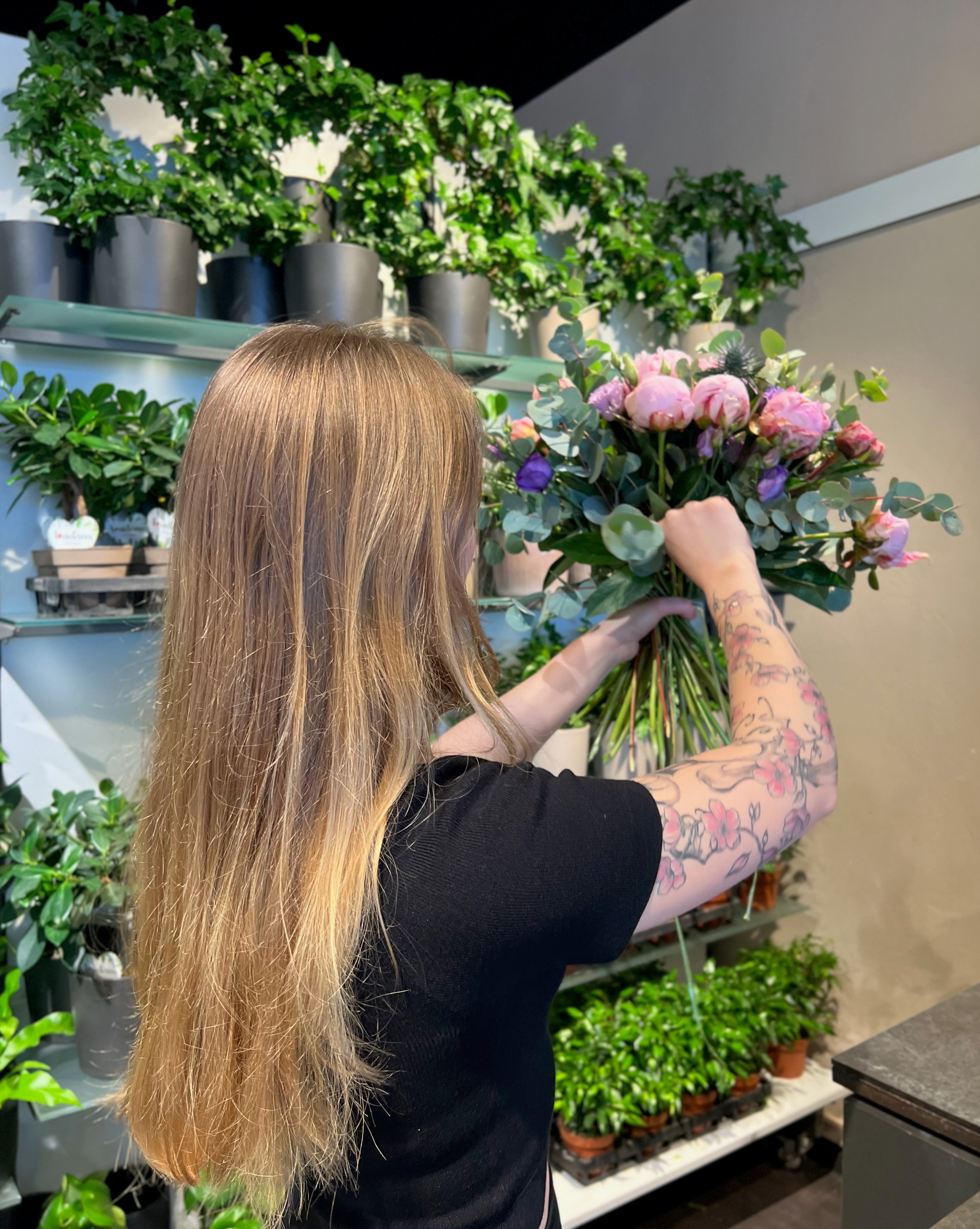 Eine Frau mit langem blondem Haar und einer Blumentätowierung arrangiert einen Strauß rosafarbener Blumen in einem Geschäft, das mit grünen Topfpflanzen und Blattranken in Regalen gefüllt ist.