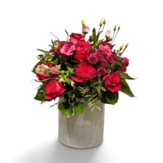 Die „Harmonie der Farben“ von florali zeigt rosa und rote Rosen mit verschiedenen Blumen und üppigem Grün, gekonnt arrangiert in einer schlichten weißen Keramikvase vor einem schlichten weißen Hintergrund.