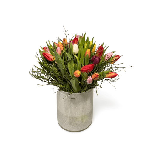 Der Tulpenstrauß „Tulip Rainbow“ von florali präsentiert bunte Tulpen in Rot, Gelb, Orange, Pink und Weiß mit Eukalyptus und Zweigen in einer Silbervase – ein frischer Farbakzent für Ihr Zuhause.