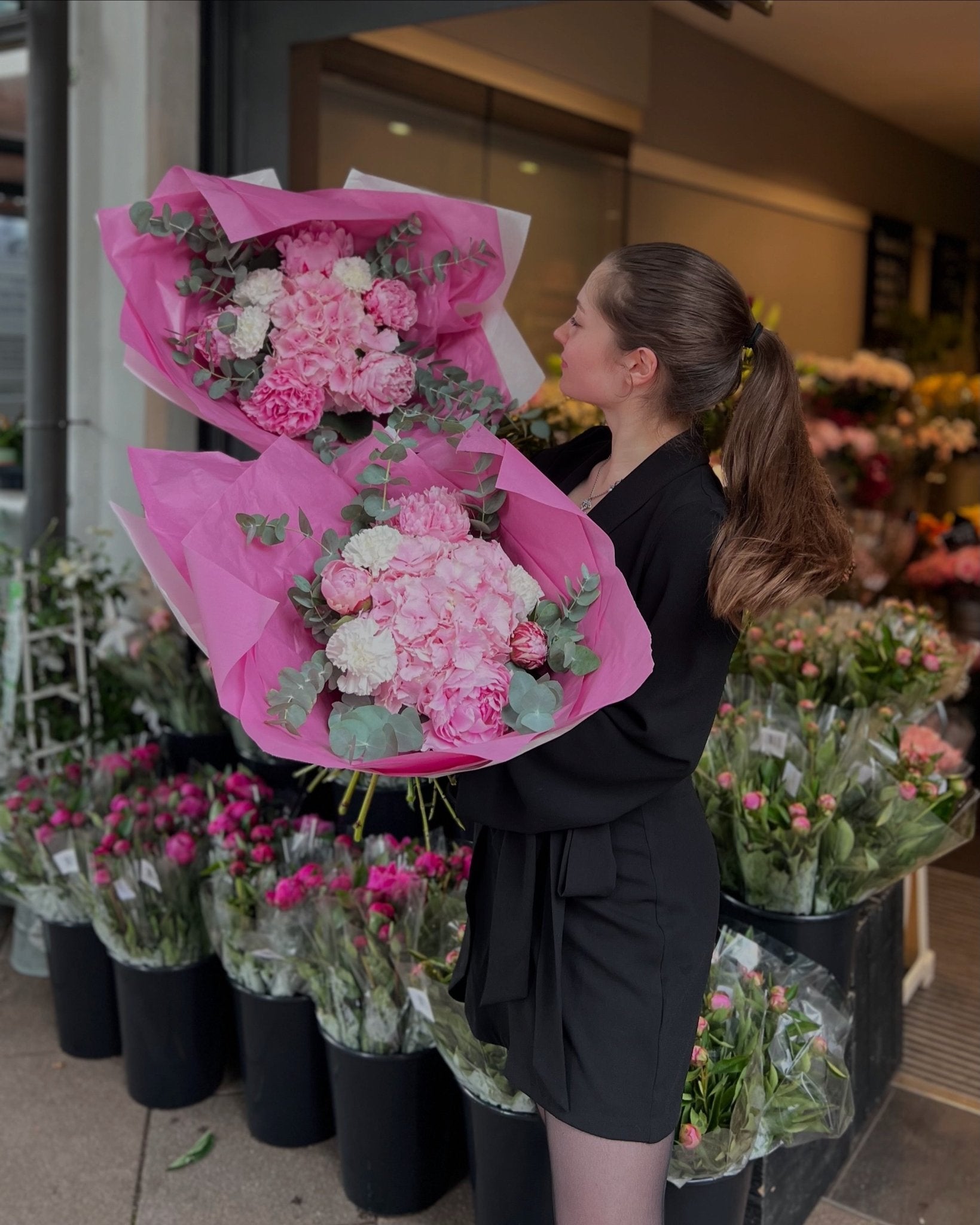 Eine junge Frau mit braunen Haaren steht vor einem offenen Blumenladen mit zwei Blumensträußen in der linken und rechten Hand. Beide Blumensträuße beinhalten Pfingstrosen, Eucalypthus und Hortensie. Im Hintergrund befinden sich Pfingstrosen in pink und rosa.