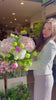 Floristin die einen Blumenstrauß mit rosa Hortensien, Schneeball und Pfingstrosen vor einem offenen Blumenladen trägt.